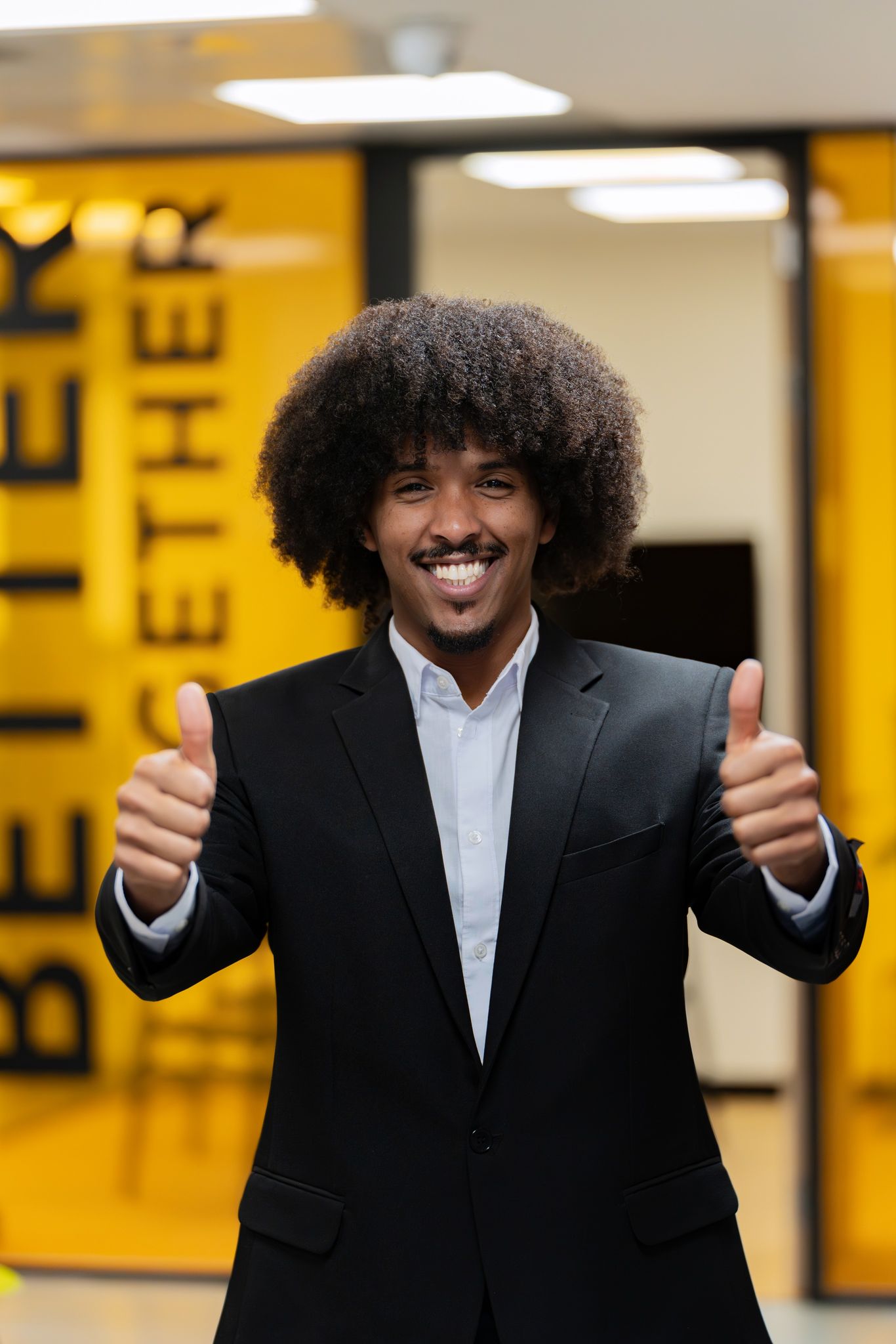 Enthusiastic businessman with afro hairstyle gives a double thumbs up in a vibrant office space.
